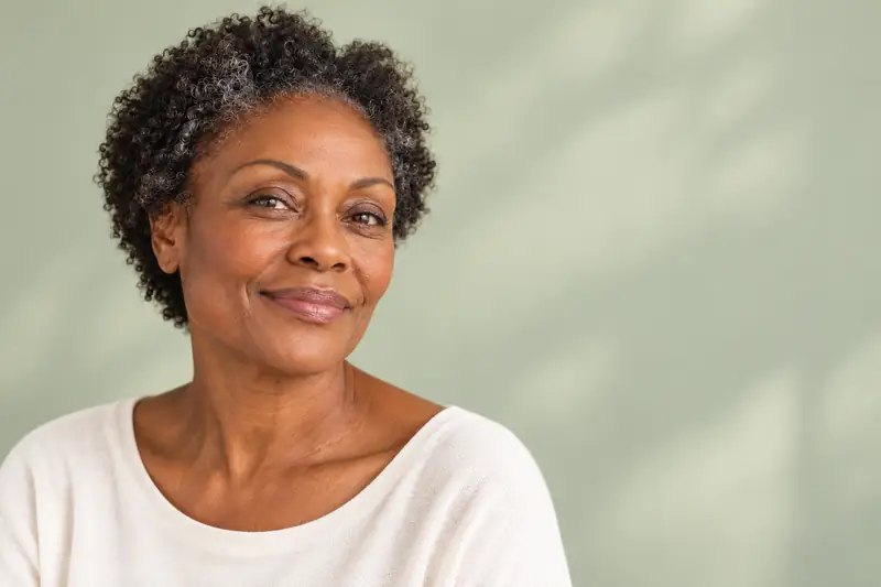 A woman in her fifties with short grey-black curls, smiling in soft daylight. Illustrative image for a skin hydrator treatment at LUMEN, Birmingham.